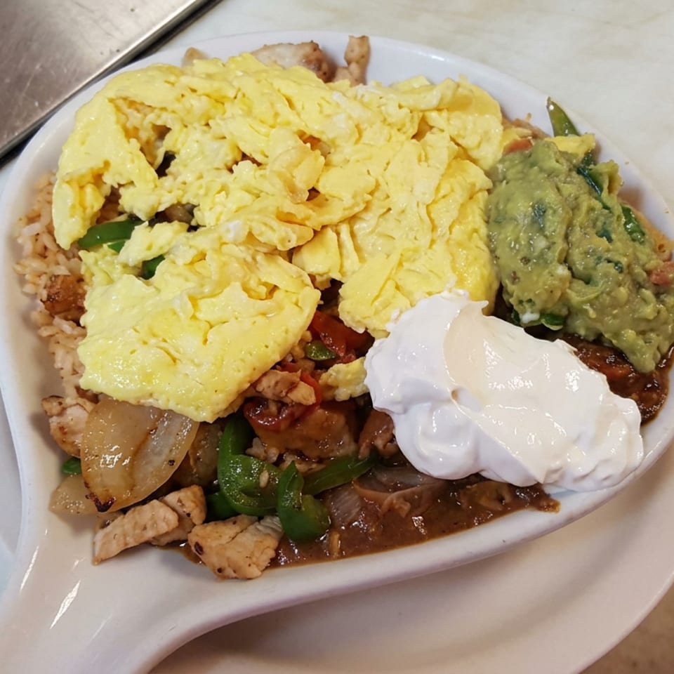 Loaded plate of rice, scrambled eggs, sautéed vegetables, meat, guacamole, sour cream, and brown sauce on a white dish.
